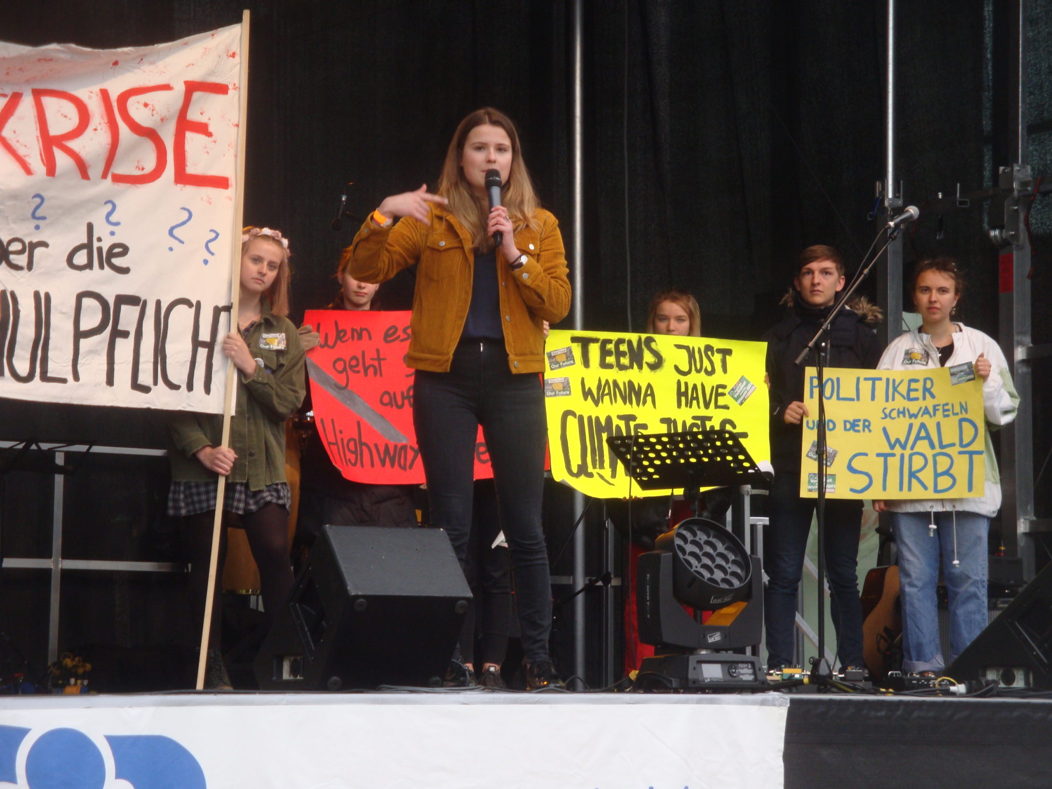 Luisa Neubauer steht auf einer Bühne mit einem Mikrofon, hinter ihr stehen andere Jugendliche mit Plakaten