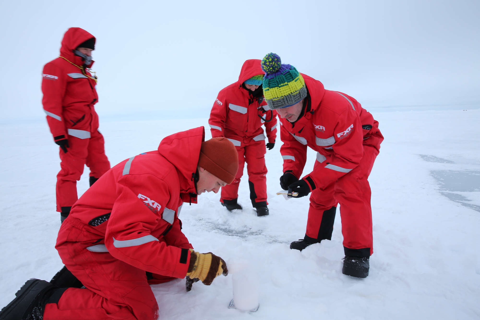Vier Forscher:innen vom AWI in roten Anzügen knien im Eis und untersuchen es
