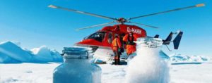 Ein roter Helikopter im arktischen Eis. Es ist strahlend blauer Himmel, im Hintergrund türmen sich Eisberge. Zwei Forscher mit dicken orangenen Überlebensanzügen stehen am Helikopter. Im Vordergrund sieht man zwei Weckgläser mit Schneeproben.