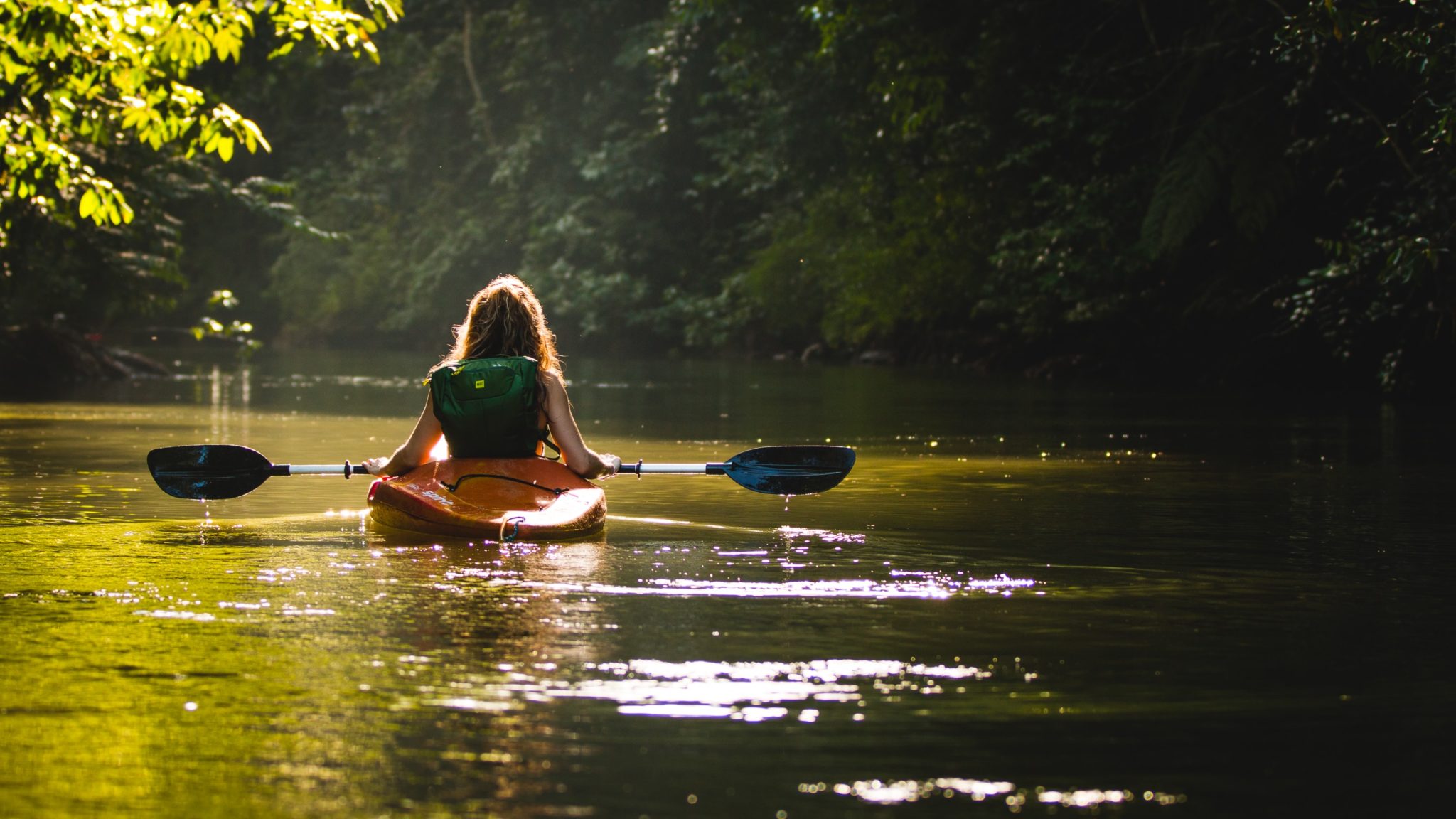 Eine Frau fährt mit einem Kanu durch einen Fluss, an den Flussufern wächst viel Grün.