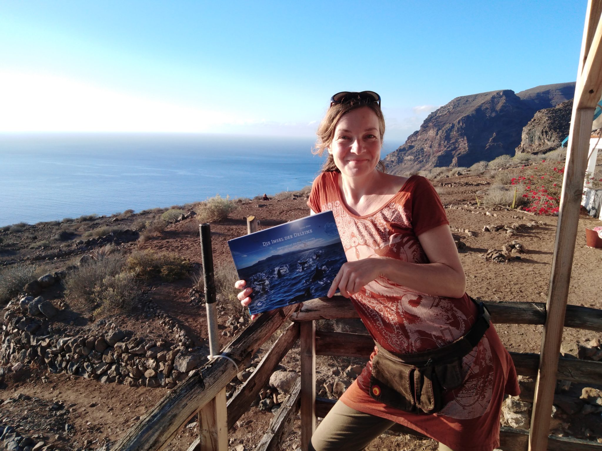 Barbara Focke steht an der Ecke der kleinen Aussichtsplattform. In ihren Händen hält sie das Buch "Die Insel der Delfine". Im Hintergrund sieht man wieder das Meer und die Berge von La Gomera