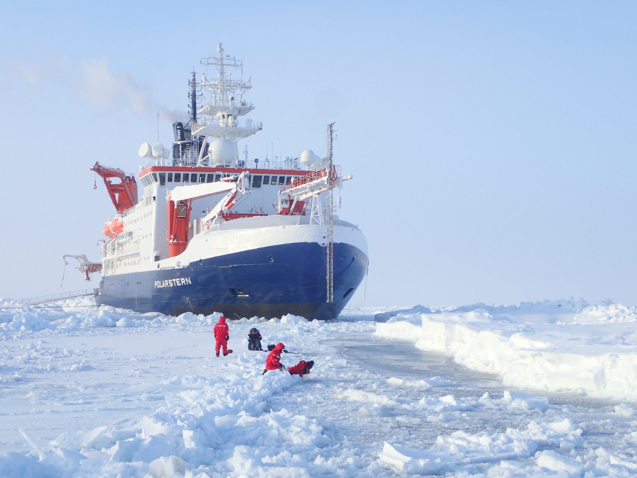 Ein Forscher steht mit rotem Anzug mitten im Eis vor dem großen Schiff Polarstern