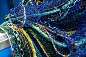 Ein mehrfarbiges Schleppnetz hängt über die Reling eines blauen Schiffes. Mehrere Seile, Tampen und Ketten liegen übereinander