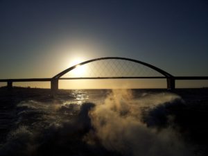 Vor der untergehenden Sonne sieht man die Brücke nach Fehmarn, im Vordergrund Wellen