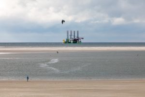 An einem Nordseestrand steht ein Kitesurfer, im Hintergrund sieht man eine Bohrinsel im Watt.