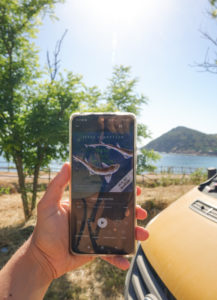 Smartphone Bildschirm mit dem Hörbuch Titel "Wenn Haie leuchten" von Julia Schnetzer drauf. Im Hintergrund sardischer Strand und Berg.