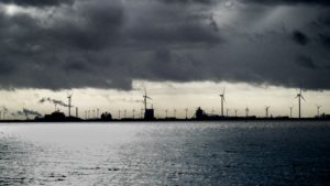 Industrie und mehrere Windräder stehen im Hintergrund im Meer. Darüber ist es stark bewölkt, die Stimmung ist düster