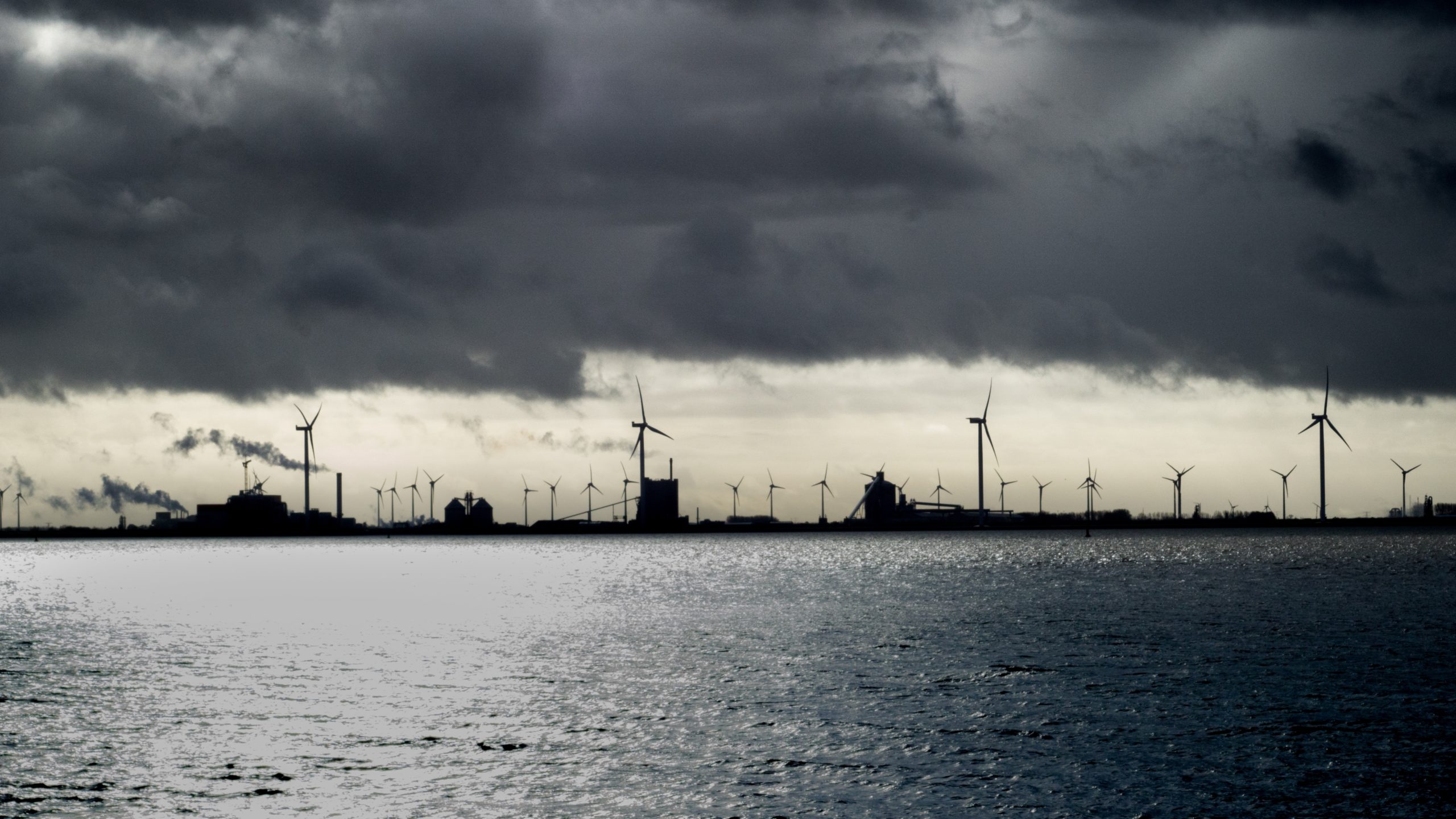 Industrie und mehrere Windräder stehen im Hintergrund im Meer. Darüber ist es stark bewölkt, die Stimmung ist düster